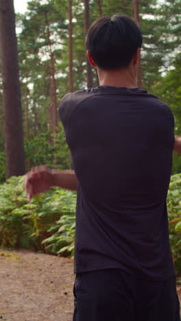 Vertical-Video-Rear-View-Shot-Of-Man-Warming-Up-And-Stretching-Arms-Before-Work-Out-Exercising-Running-Through-Forest-1